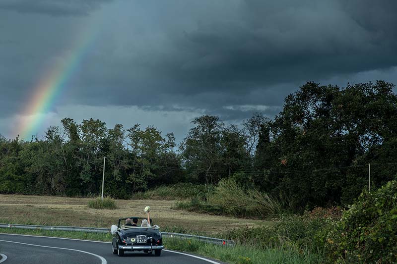 Un arcobaleno illumina il cielo cupo mentre sulla via corre l'auto degli sposi